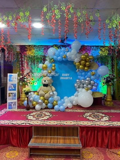 A full view of the banquet hall stage decorated for a baby's party. The setup includes a teddy bear theme, 'BABY' blocks, and floral arrangements on the side.