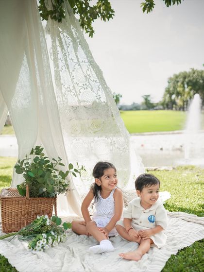 Two siblings sit together on a picnic blanket, smiling and enjoying the beautiful day. The fountain in the background adds a lovely touch to the scene.
