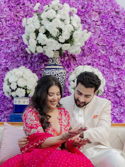 The groom admires the bride's intricate henna design during their Mehendi. This candid moment is full of admiration and love, set against a stunning purple and white floral wall.