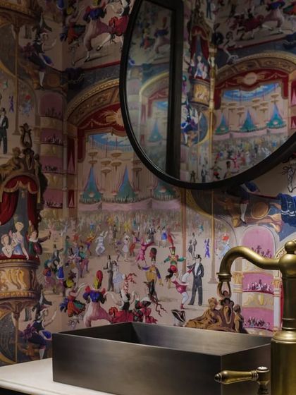 The powder room in the Caitriona apartment is a moment of pure whimsy and design wit. The walls are covered in a vibrant 'Cole & Sons' wallpaper depicting cabaret dancers, turning a small space into a memorable and playful experience.
