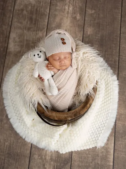 A sleeping newborn snuggles with a tiny teddy bear. This overhead shot in a basket shows the baby's peaceful expression and the cozy, neutral-toned setup.