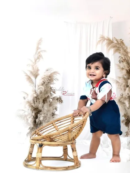 A milestone photo capturing a baby boy proudly standing with the help of a small wicker chair. The minimalist, boho-style setup with pampas grass is very modern and stylish.