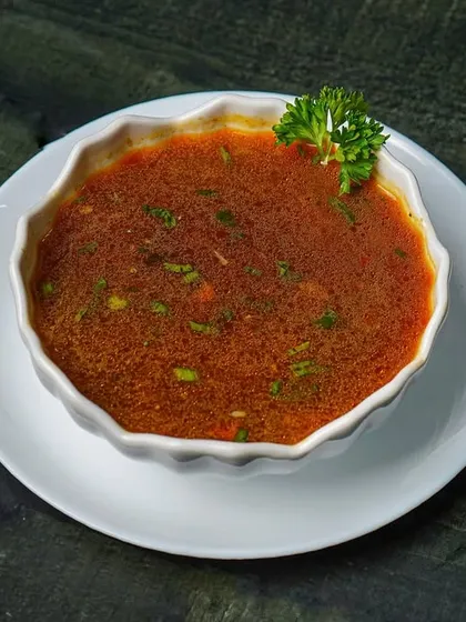 A bowl of hearty tomato soup garnished with parsley, shot from above on a dark wooden table. The deep red of the soup contrasts beautifully with the dark background.