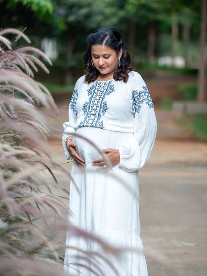 A serene outdoor portrait of a mom-to-be in a white embroidered dress, cradling her bump amidst tall, soft grasses.