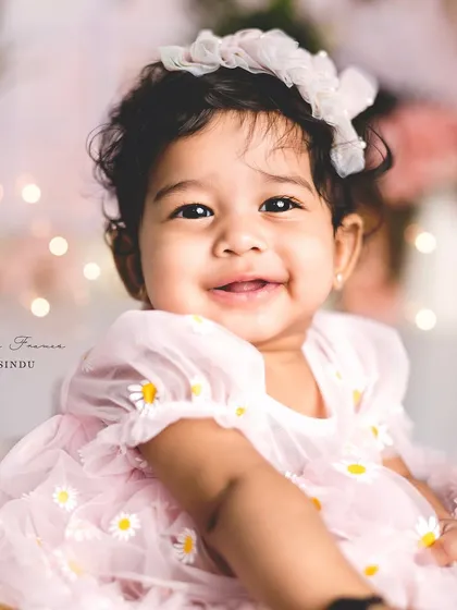 A close-up of a happy baby girl in a pink daisy dress. The bokeh lights in the background add a touch of magic to this sitter portrait.