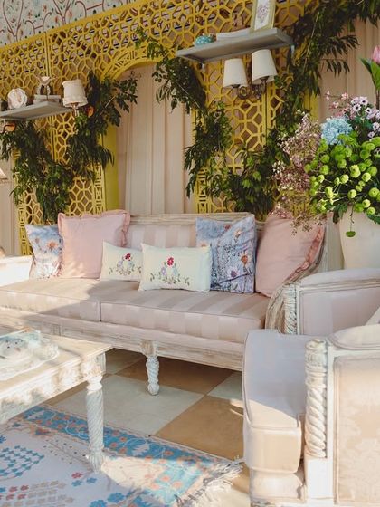 A beautiful lounge area from the pastel Mehendi. The decor featured custom floral-print cushions, distressed white furniture, and a yellow jaali backdrop, creating a chic and comfortable space for guests.