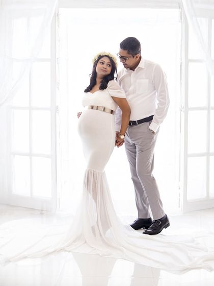 A classic and elegant couple's portrait against a bright, backlit window. The mom-to-be wears a simple white gown, creating a timeless and sophisticated look.