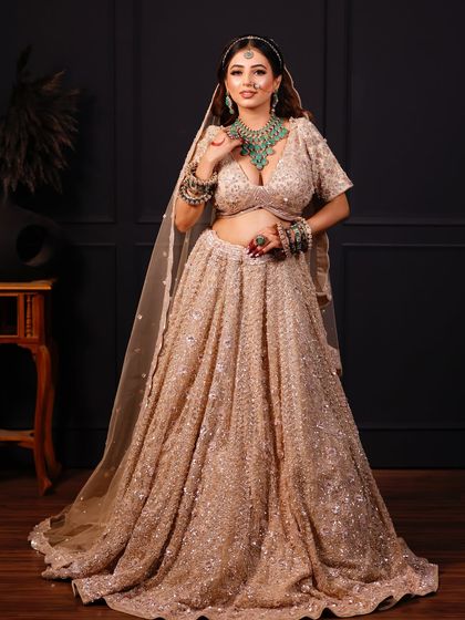 A full-length shot of a bride in a sparkling champagne-colored lehenga, paired with a contrasting green necklace.
