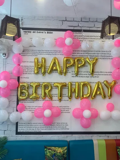 A birthday decoration at a restaurant, featuring 'Happy Birthday' foil balloons framed by pink and white balloon flowers against a white brick wall.