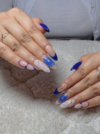 A striking design with a bold blue French tip, a sheer nude base, and a white floral pattern with blue and gold accents.