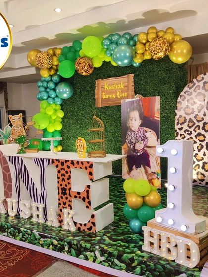 A wide shot of a jungle-themed first birthday. This setup includes a green wall backdrop, a 'ONE' marquee table, and multiple animal character cutouts.