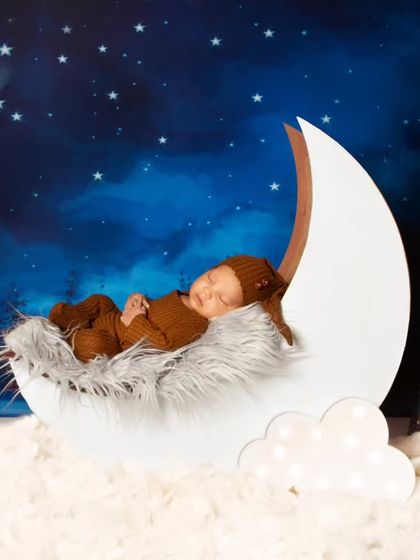 A newborn in a cozy brown outfit sleeps peacefully inside a white crescent moon prop. This setup uses a softer, fluffier texture for the moon and clouds.