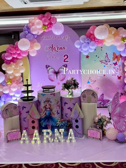 A magical butterfly theme for a second birthday. This elaborate setup features large butterfly wing props, themed pedestals, and a pastel balloon arch against a purple backdrop.