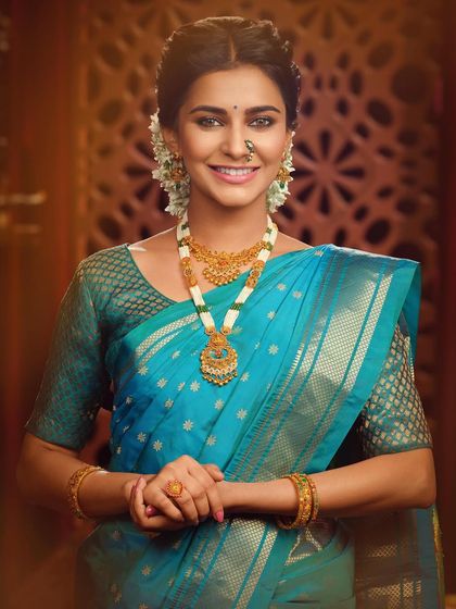 A smiling portrait highlighting a different set of traditional jewelry. The pearl and gold necklace adds a touch of softness to the vibrant blue saree, creating a look of pure elegance.