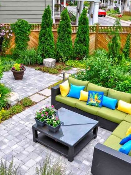 This image shows a beautifully executed small backyard. The design uses a paved patio for seating, surrounded by multi-level garden beds filled with a variety of shrubs, flowers, and trees to create a sense of privacy and enclosure.
