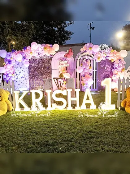 An elaborate outdoor butterfly-themed stage for Krisha's first birthday. The setup on the lawn includes marquee letters, large teddy bears, and multiple backdrops with balloon arches.
