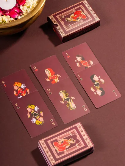 A styled shot of Namostutay's traditionally illustrated playing cards. The rich, warm tones and festive props like a brass bowl of flowers create a cultural and celebratory context.