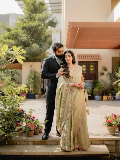 An engagement portrait that exudes grace and sophistication. The groom gently holds his bride, both dressed in elegant attire, surrounded by the lush greenery of a beautiful home garden.
