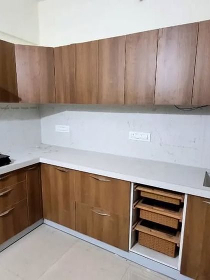 A classic L-shaped modular kitchen with a warm wooden laminate finish. This design is timeless, offering ample storage and counter space with an integrated vegetable basket pull-out.