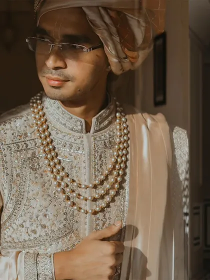 A classic, elegant portrait of a groom in his sherwani and pearl necklace, with soft window light highlighting his features.