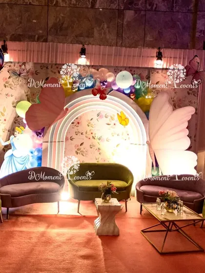 A cozy and elegant seating area within a princess and butterfly-themed party. The backdrop features a floral pattern, large butterfly wings, and a cutout of Cinderella, with comfortable sofas for guests.