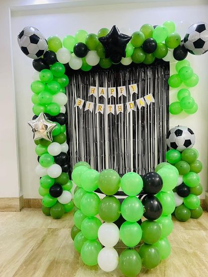 A sporty football-themed birthday decoration. The setup features a goal-like backdrop with a black shimmer curtain and a balloon frame in green, black, and white, accented with football-shaped foil balloons.