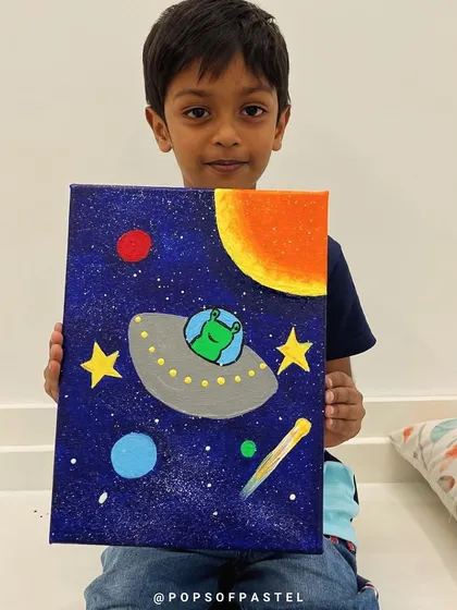 A young boy proudly holds up his out-of-this-world space-themed acrylic painting, created with guidance from our amazing teacher.