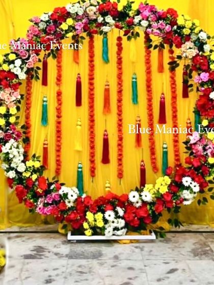 A beautiful Mehendi backdrop featuring a full ring of colorful flowers and hanging tassels against a bright yellow background.