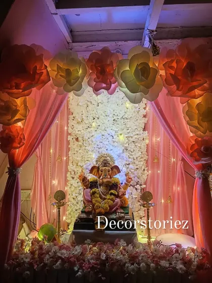 We designed this grand and elegant home setup for the Ganpati festival. The backdrop features a wall of white artificial flowers, soft peach draping, and oversized paper flowers, all illuminated to create a divine atmosphere for Bappa.