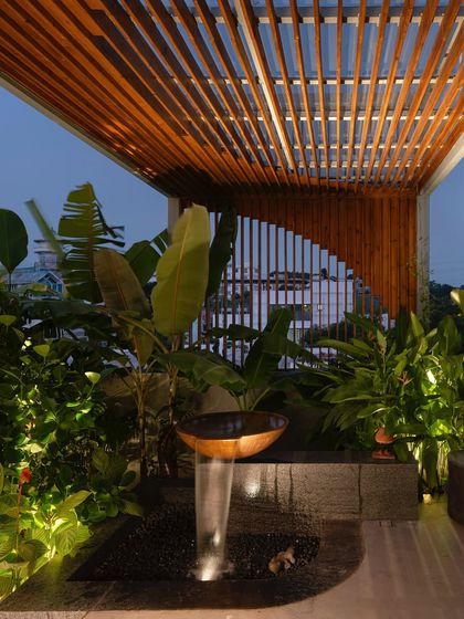 A close-up of a rooftop water feature at dusk, with a wooden pergola overhead. The combination of flowing water, warm light, and tropical plants creates a serene and inviting sensory experience.