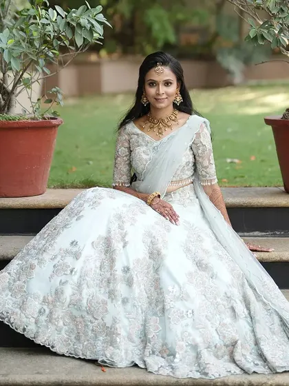 A bride seated in her ethereal light blue lehenga. The entire piece is covered in delicate white floral embroidery, creating a dreamy and romantic look for a daytime wedding or reception.