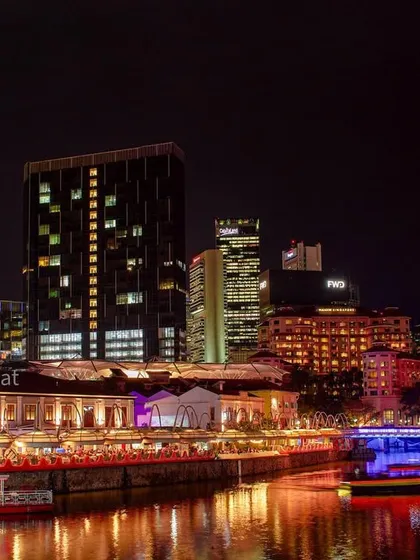 A night view of Clarke Quay in Singapore, showing the illuminated restaurants and their reflections on the river.