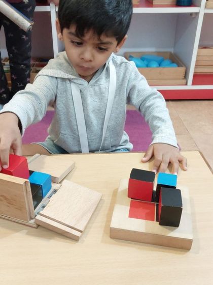 A child works to solve the puzzle of the Binomial Cube. The material is self-correcting; if the pieces don't form a perfect cube that fits in the box, the child knows to try again, fostering independent problem-solving.