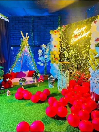 An elaborate indoor-outdoor themed romantic setup. It features a canopy tent on artificial grass, a shimmering backdrop, and plenty of red balloons to set a passionate mood.