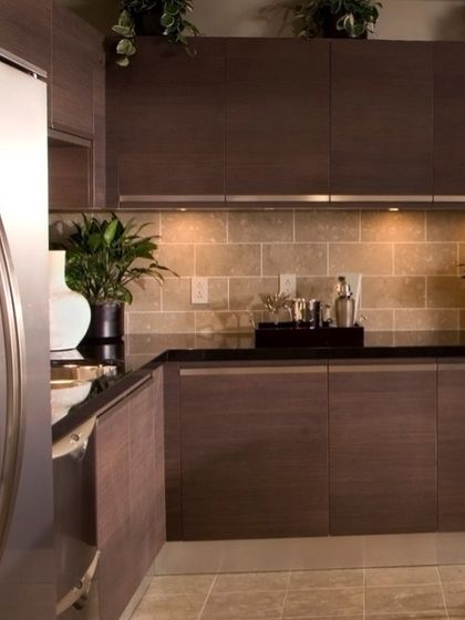 A close-up of a kitchen with dark wood-grain cabinets and a black countertop. The under-cabinet lighting highlights the tiled backsplash and creates a warm, intimate mood.