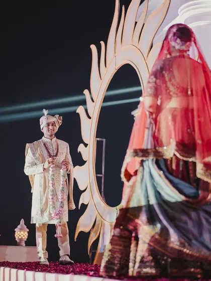 A moment of anticipation before the union. The groom waits with the garland as the bride, seen through a beautiful sheer veil, approaches for the varmala.