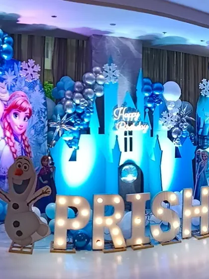 Bringing the magic of Arendelle to life with a complete Frozen theme party setup. This includes character cutouts of Elsa, Anna, and Olaf, an illuminated ice castle backdrop, snowflake details, and a balloon garland in signature icy blue, white, and silver tones. The light-up marquee letters for the name make it extra special.
