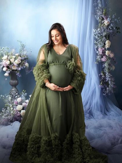 A full-length view of the green gown against a soft blue floral background. The mom-to-be cradles her bump, looking serene and beautiful.
