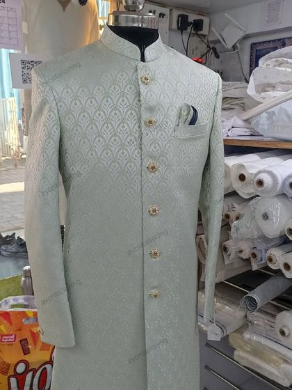 A full view of a custom-stitched wedding sherwani in a light, refreshing color. This piece is displayed in my store, surrounded by rolls of premium fabric, showing the possibilities for your own custom creation.