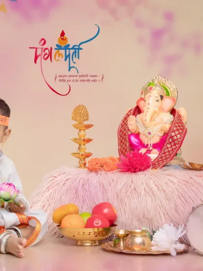 A smiling baby boy in a white kurta with a "Ganpati Bappa Morya" headband. The festive setup includes a dholak, a lotus flower, and an oil lamp, perfect for a milestone shoot during Ganesh Chaturthi.