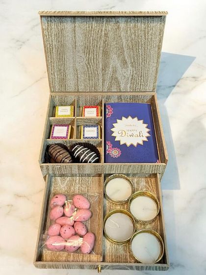 A luxurious two-tiered wooden box for Diwali. The top layer holds assorted chocolates and a greeting card, while the bottom drawer reveals scented candles and sweets.