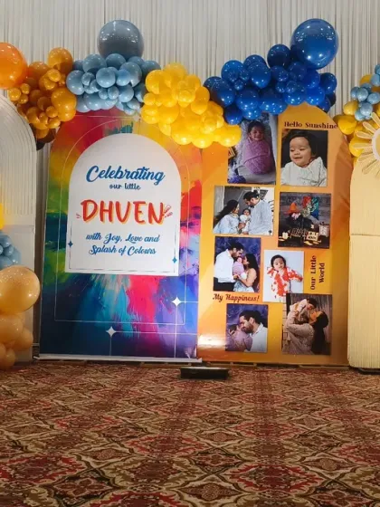 A colorful and joyful first birthday backdrop featuring a photo collage. The theme is 'A Splash of Colors', reflected in the vibrant balloons and rainbow-splashed design.