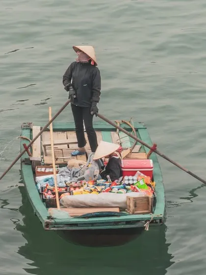 A vendor stands to paddle their floating shop, filled with goods, across the calm waters of Halong Bay.