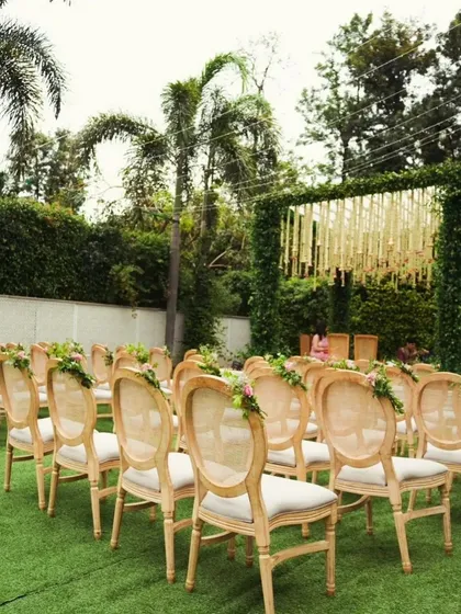 An outdoor ceremony seating arrangement with elegant wooden chairs on a lush green lawn. The aisle is clear, leading to a beautiful mandap covered in greenery.
