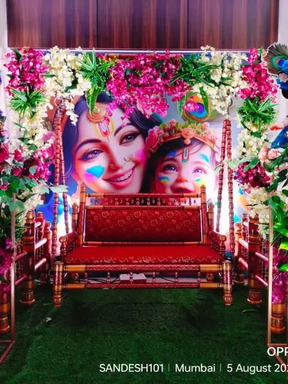A close-up of a Krishna-themed jhula decoration for a naming ceremony. The cradle is placed in front of a vibrant backdrop and surrounded by floral arrangements.