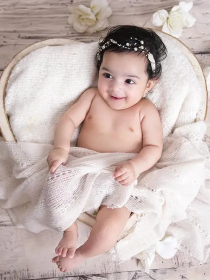 A happy baby girl with a pearl headband, sitting up in a white basket surrounded by flowers. Her smile is absolutely radiant.