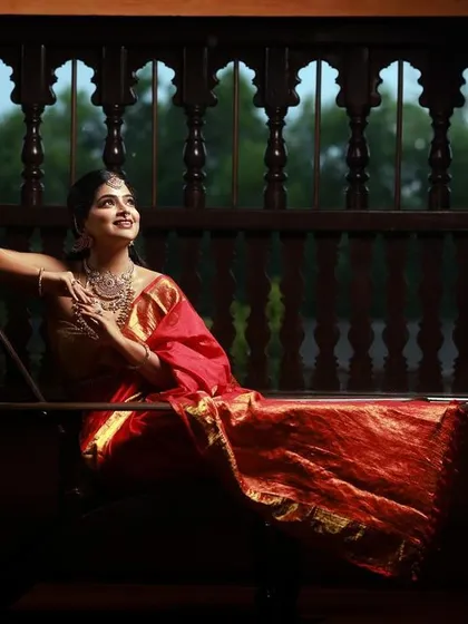 The intricate wooden screen provides a beautiful, textured backdrop for this portrait. The low-angle shot emphasizes the bride's regal posture and the flowing drape of her red saree.
