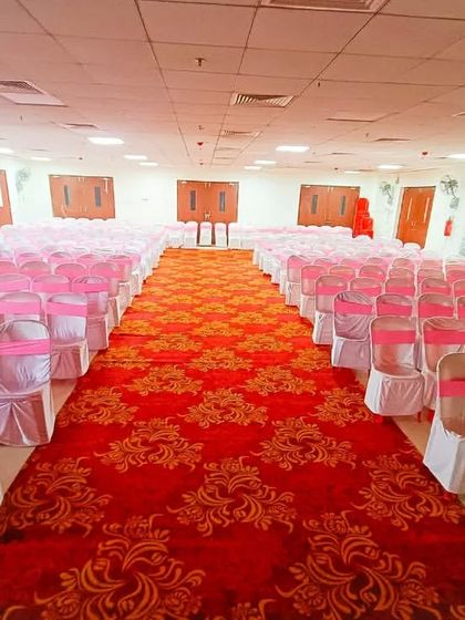 I can handle the entire venue setup, including seating arrangements. This image shows a hall prepared for a large ceremony with chairs neatly arranged with pink sashes and a red carpet aisle.