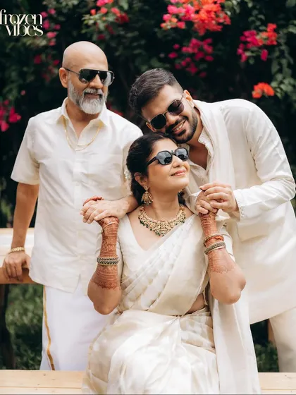 A modern family portrait, captured outdoors with natural light. The son embraces his mother, who is seated, while the father stands beside them, all sharing a moment of relaxed connection in coordinated white outfits.
