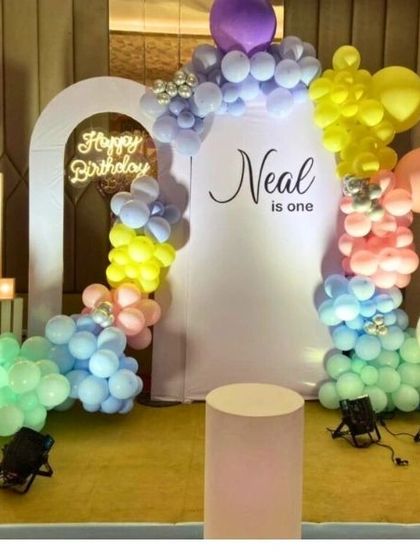 A colorful and cheerful first birthday for "Neal." This setup uses a pastel rainbow balloon arch against a white backdrop, a "Happy Birthday" neon sign, and a light-up number "1."